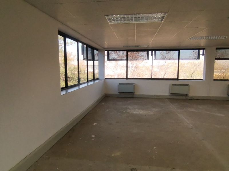 An empty, open-plan commercial office space featuring large windows, wall-mounted HVAC units, and a suspended ceiling with recessed lighting.