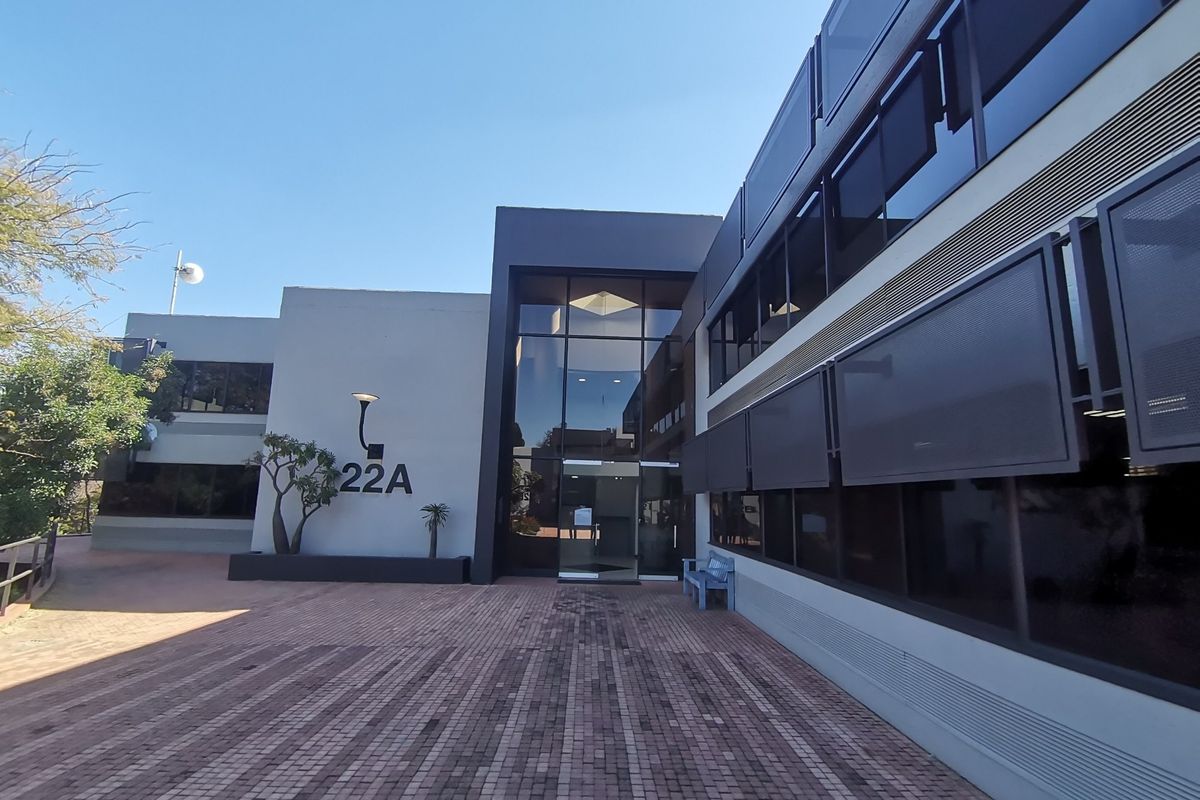 Modern commercial building exterior featuring a glass entrance facade, paved forecourt, and integrated external sun shading systems.