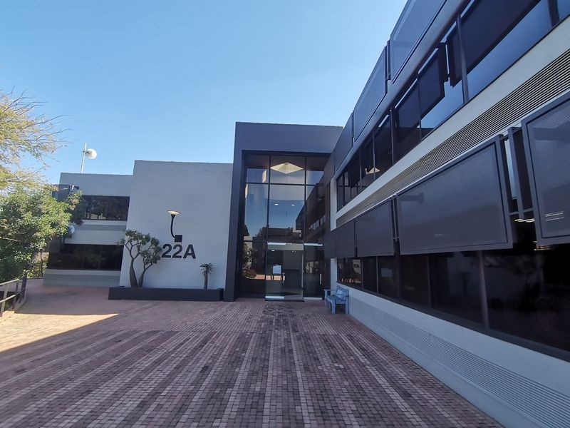 Modern commercial building exterior featuring a glass entrance facade, paved forecourt, and integrated external sun shading systems.