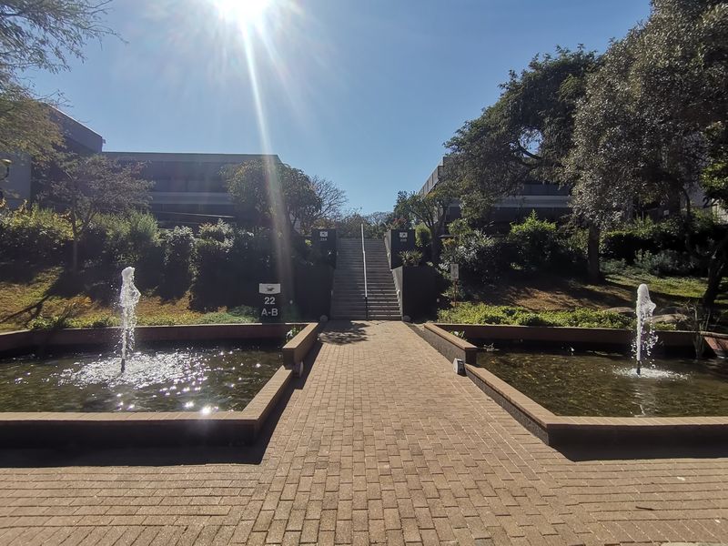 A well-maintained commercial office park entrance featuring a paved walkway, central water fountains, and concrete stairs leading to the building complex.