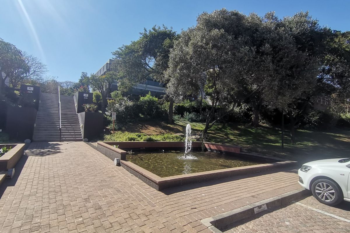 Exterior view of a commercial property featuring a landscaped garden, a central water fountain, and a paved pedestrian walkway leading to a staircase.