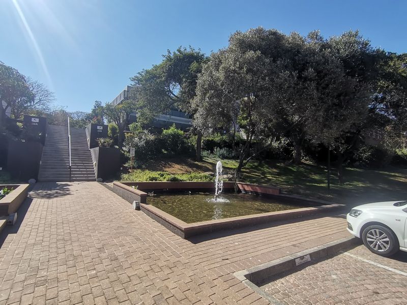 Exterior view of a commercial property featuring a landscaped garden, a central water fountain, and a paved pedestrian walkway leading to a staircase.