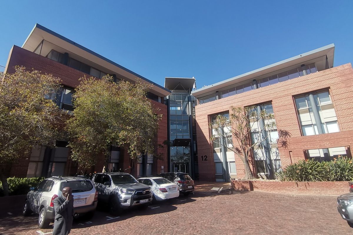 Front elevation of a multi-story commercial office building featuring a red brick facade, glass entrance atrium, and a paved parking area.