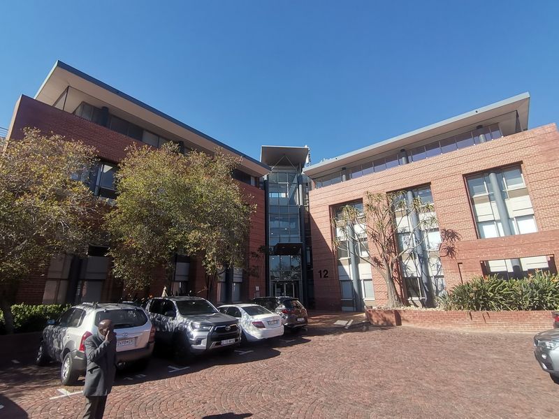 Front elevation of a multi-story commercial office building featuring a red brick facade, glass entrance atrium, and a paved parking area.