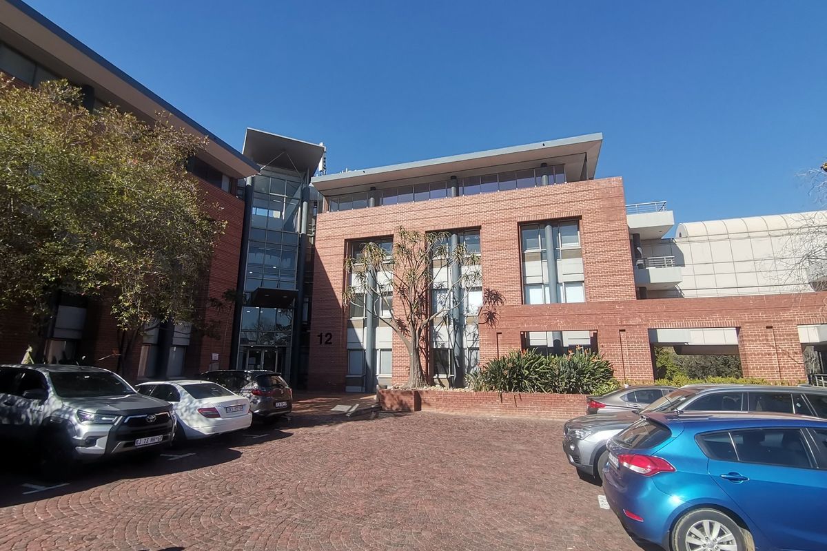 Front elevation of a multi-story commercial office building featuring a brick facade, glass entrance section, and a paved parking area.
