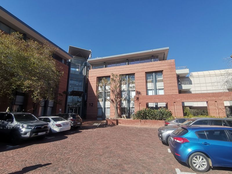 Front elevation of a multi-story commercial office building featuring a brick facade, glass entrance section, and a paved parking area.