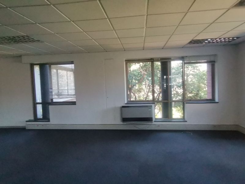 An empty, open-plan office space featuring a suspended ceiling, wall-mounted air conditioning unit, and large windows providing natural light.