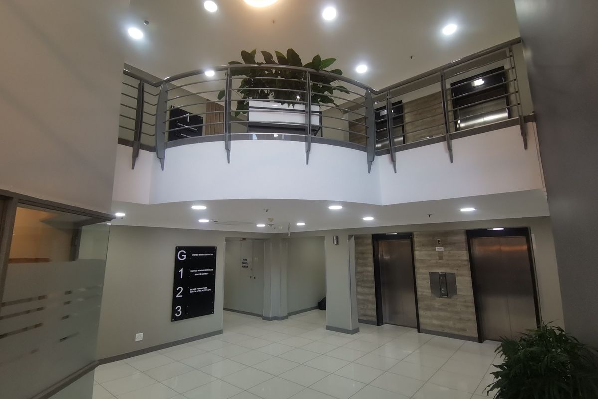 Modern commercial building lobby featuring a double-volume entrance, mezzanine level with metal balustrade, tiled flooring, and elevator access.