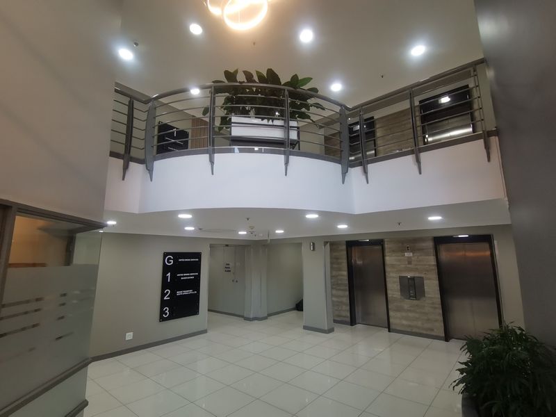 Modern commercial building lobby featuring a double-volume entrance, mezzanine level with metal balustrade, tiled flooring, and elevator access.