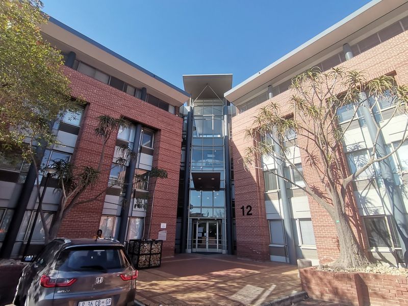 Front elevation of a modern commercial office building featuring a prominent glass entrance atrium, red brick facade, and paved approach.