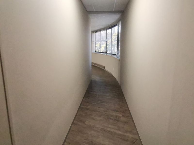 A curved commercial hallway featuring neutral walls, laminate flooring, and large windows providing natural light.