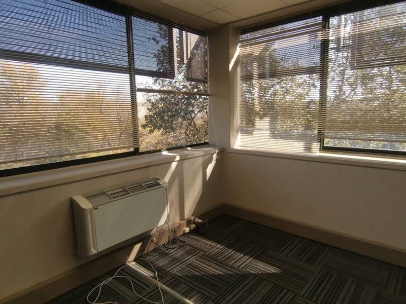 A vacant corner office space featuring large windows with venetian blinds, wall-mounted HVAC unit, and carpet tile flooring.