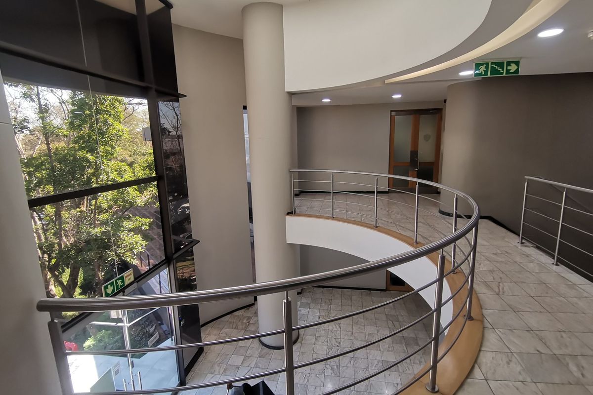 Modern commercial building lobby featuring a curved staircase with stainless steel balustrades, marble flooring, and expansive floor-to-ceiling windows providing natural light.