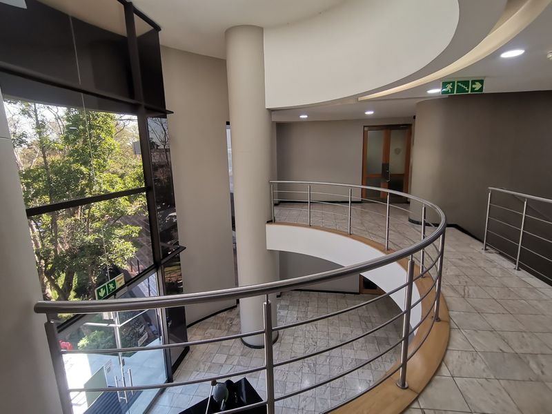 Modern commercial building lobby featuring a curved staircase with stainless steel balustrades, marble flooring, and expansive floor-to-ceiling windows providing natural light.