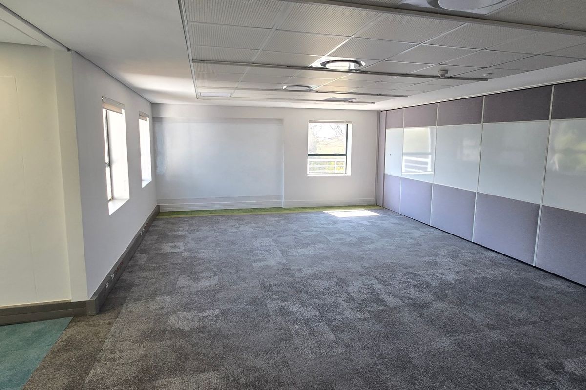 A spacious, empty commercial office area featuring carpeted flooring, a suspended ceiling with recessed lighting, and integrated whiteboard wall panels.