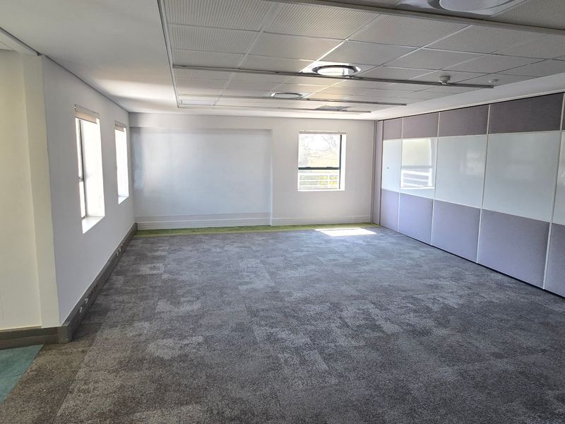 A spacious, empty commercial office area featuring carpeted flooring, a suspended ceiling with recessed lighting, and integrated whiteboard wall panels.