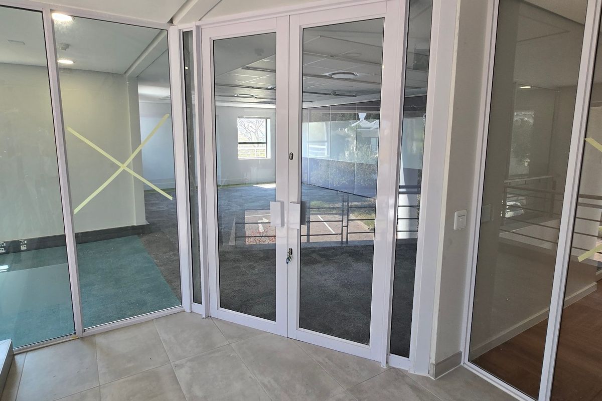 Modern commercial office space featuring glass partitioning, double glass doors, and a combination of tiled and carpeted flooring in a bright, open layout.
