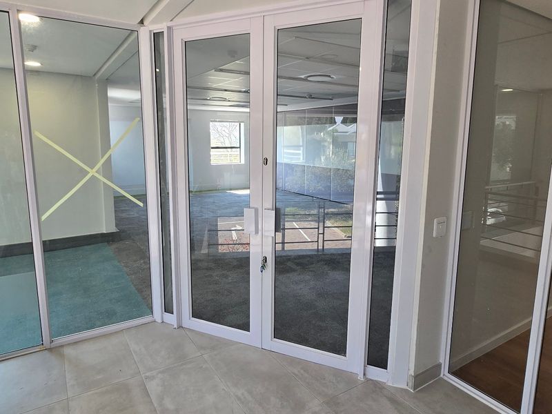 Modern commercial office space featuring glass partitioning, double glass doors, and a combination of tiled and carpeted flooring in a bright, open layout.