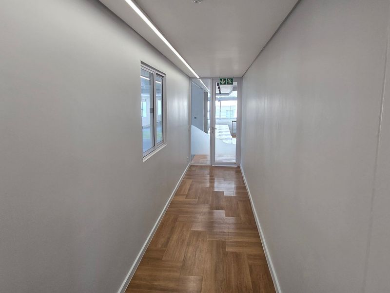 A clean, modern commercial hallway featuring herringbone wood flooring, linear ceiling lighting, and a glass partition door leading to an adjacent office space.