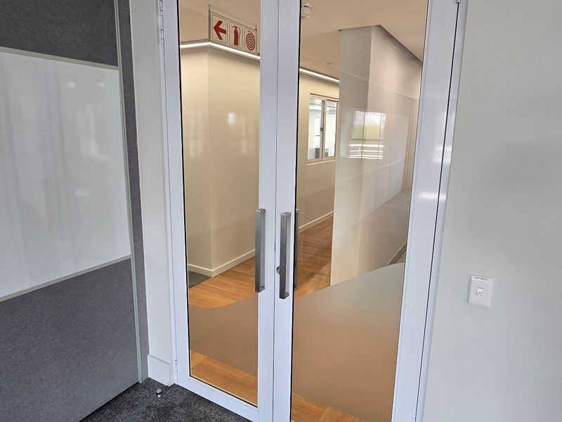 A modern commercial office entrance featuring double glass doors, clean white walls, and a combination of carpet and wood laminate flooring.