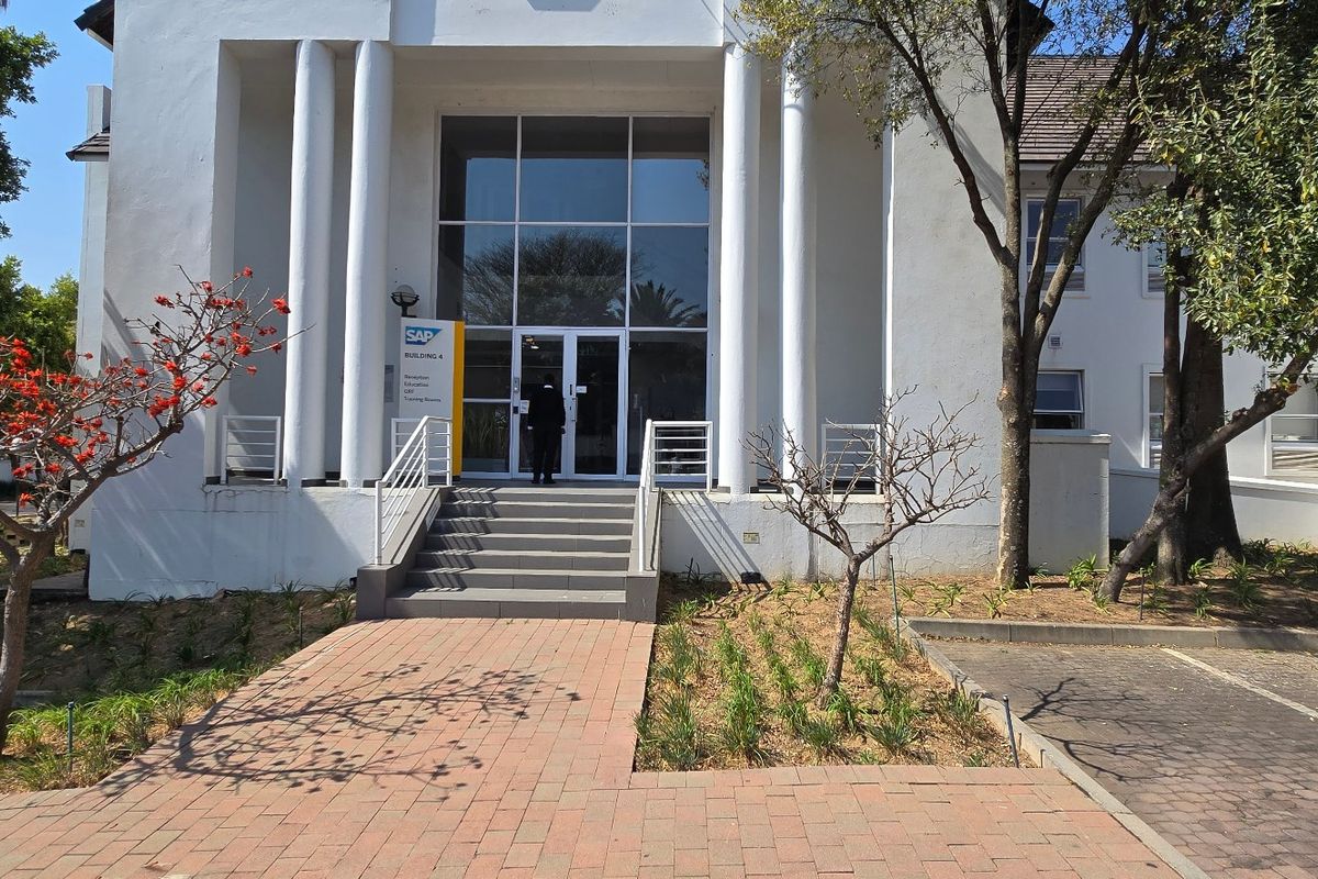 Front entrance of a commercial building featuring a glass facade, prominent concrete columns, and a paved walkway leading to the main doors.