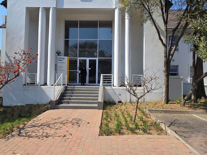 Front entrance of a commercial building featuring a glass facade, prominent concrete columns, and a paved walkway leading to the main doors.