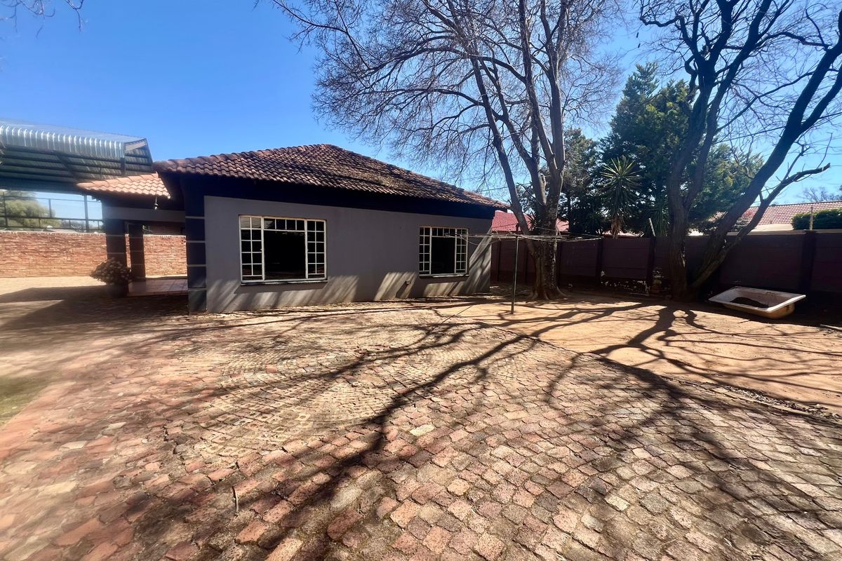 Rear exterior view of the property featuring a paved courtyard, covered patio area, and established trees within a walled boundary.