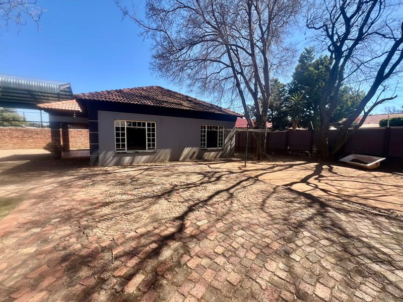 Rear exterior view of the property featuring a paved courtyard, covered patio area, and established trees within a walled boundary.