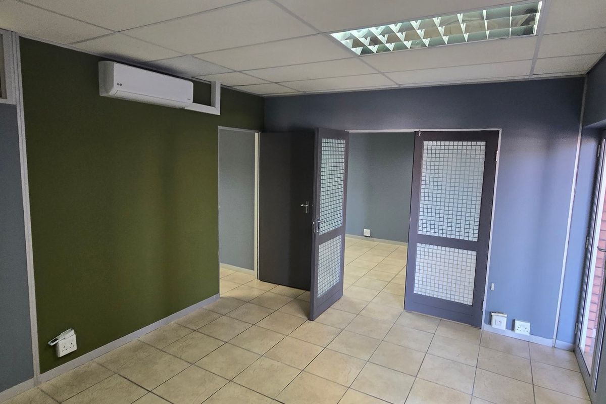 A clean, vacant office space featuring tiled flooring, a suspended ceiling with recessed lighting, and a wall-mounted air conditioning unit.