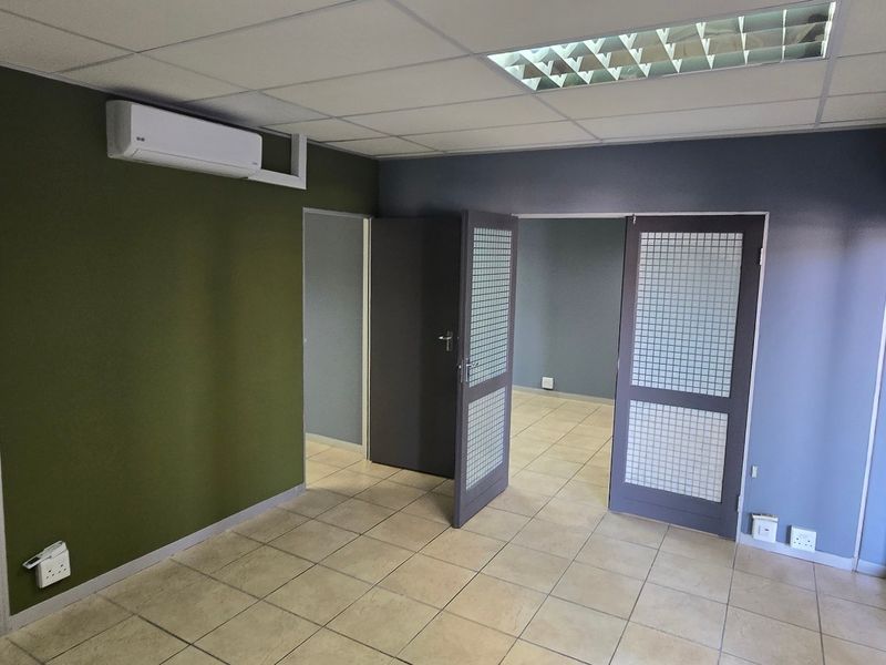 A clean, vacant office space featuring tiled flooring, a suspended ceiling with recessed lighting, and a wall-mounted air conditioning unit.