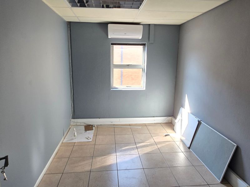 A small, empty office space featuring tiled flooring, a wall-mounted air conditioning unit, and a drop ceiling.