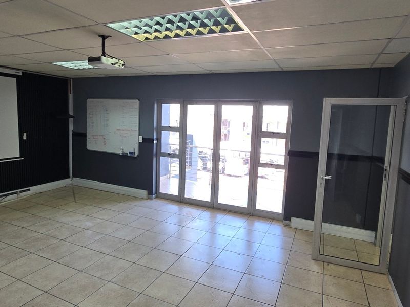 A spacious, empty office area featuring tiled flooring, a suspended ceiling with recessed lighting, a projection screen, and glass doors leading to an exterior space.