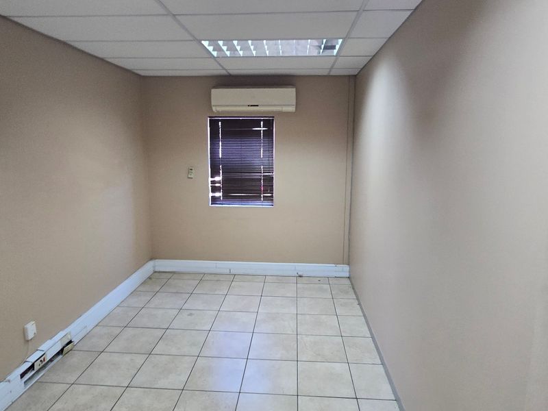 A vacant commercial office space featuring tiled flooring, a suspended ceiling with recessed lighting, and a wall-mounted split-system air conditioning unit.