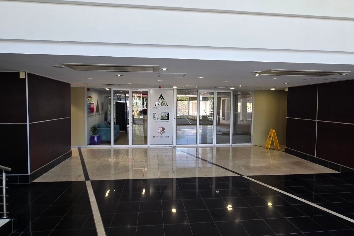 A spacious commercial building entrance lobby featuring polished tiled flooring, a modern elevator, and glass-paneled double doors leading to the main office area.