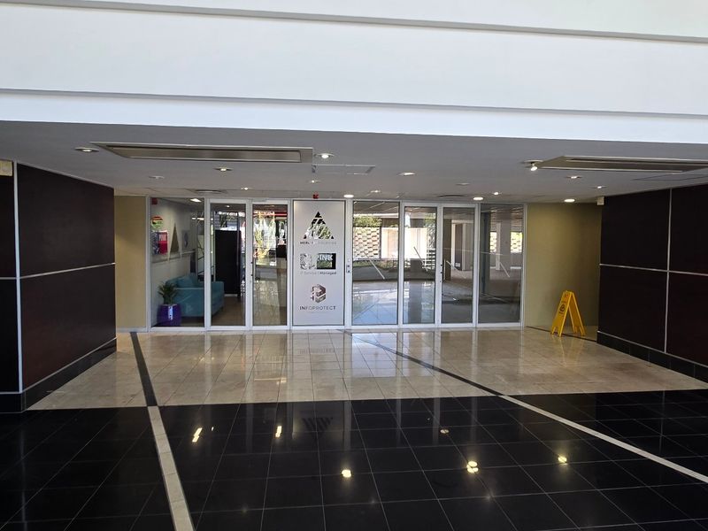A spacious commercial building entrance lobby featuring polished tiled flooring, a modern elevator, and glass-paneled double doors leading to the main office area.
