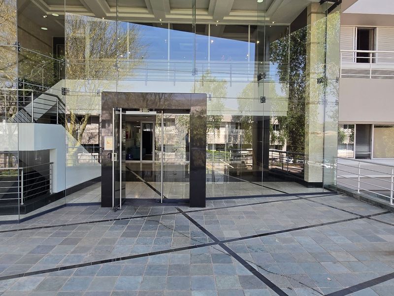 Modern commercial building entrance featuring a prominent glass facade, stone-tiled forecourt, and clean architectural lines.