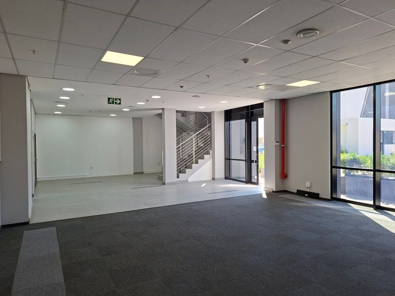 Spacious open-plan industrial office area featuring a suspended ceiling, recessed lighting, large windows, and a staircase with metal railings.
