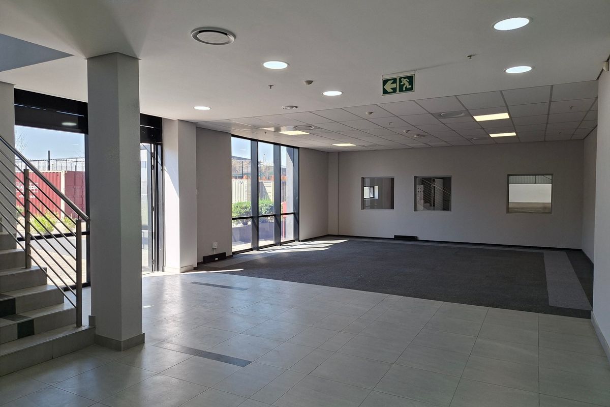 Spacious open-plan industrial office area featuring a mix of tiled and carpeted flooring, recessed lighting, and large windows providing ample natural light.