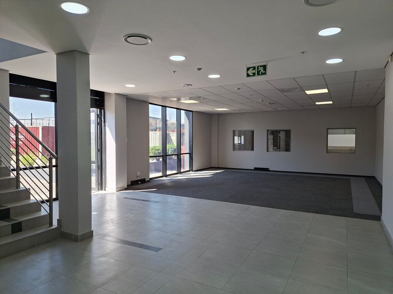 Spacious open-plan industrial office area featuring a mix of tiled and carpeted flooring, recessed lighting, and large windows providing ample natural light.