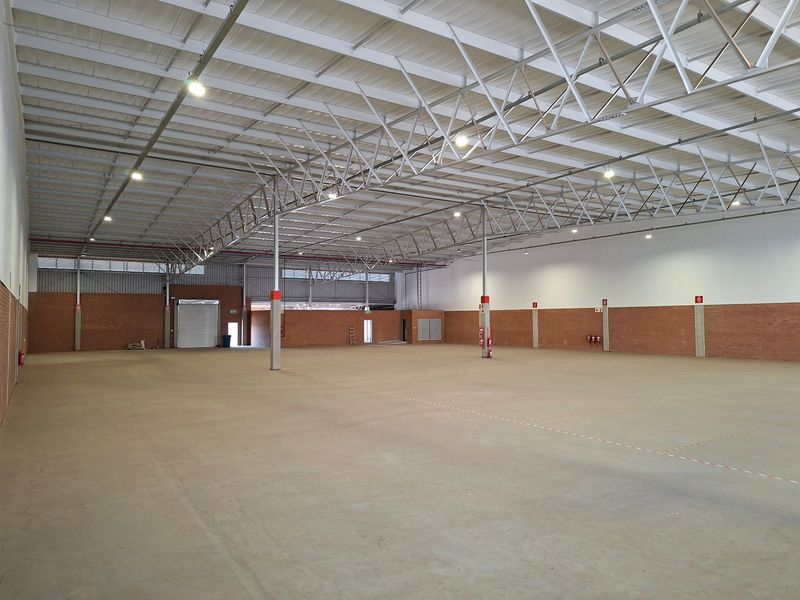 Spacious industrial warehouse featuring high ceilings with exposed steel trusses, concrete flooring, exposed brick walls, and a large roller shutter door.