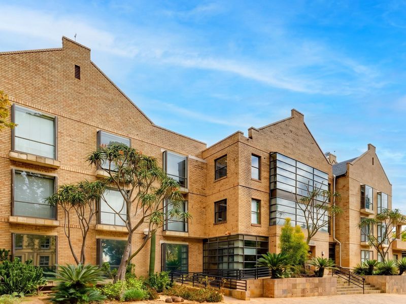 A modern multi-story commercial building featuring a distinctive brick facade, large glass windows, and well-maintained landscaped gardens with paved access.