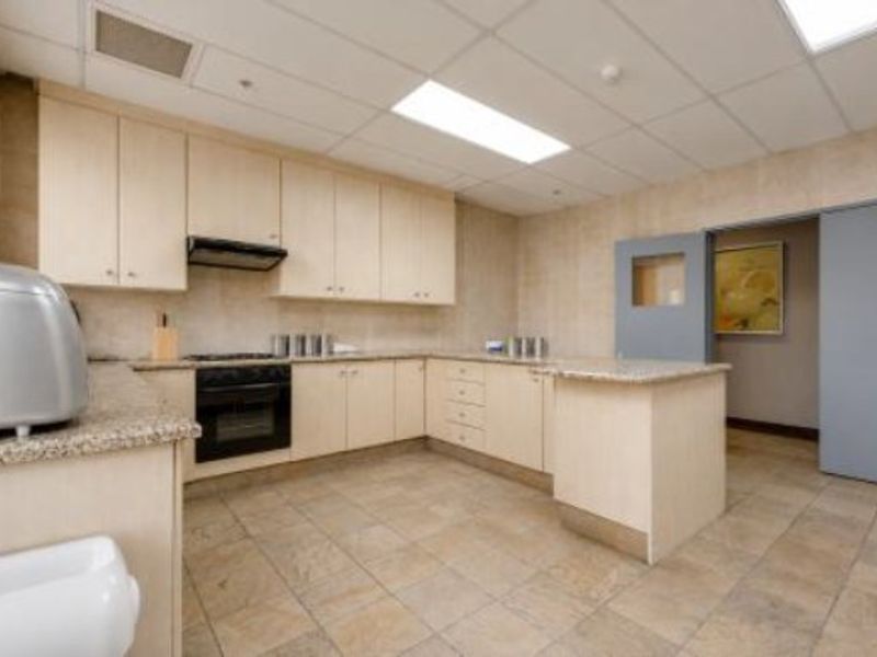 A functional commercial kitchen featuring light wood cabinetry, granite countertops, integrated oven, extractor fan, and durable tiled flooring with recessed ceiling lighting.