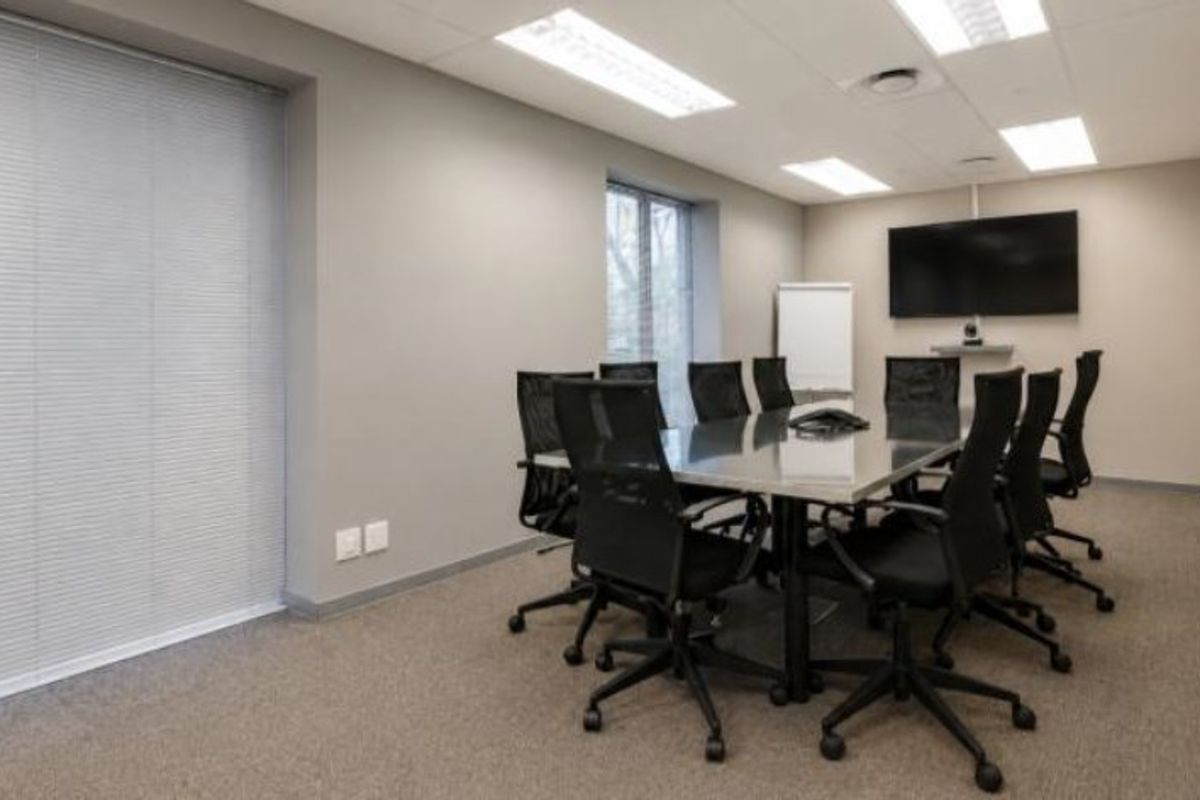 A professional commercial boardroom featuring carpeted flooring, recessed lighting, venetian blinds, and a wall-mounted television for presentations.