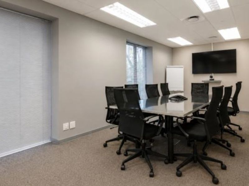 A professional commercial boardroom featuring carpeted flooring, recessed lighting, venetian blinds, and a wall-mounted television for presentations.