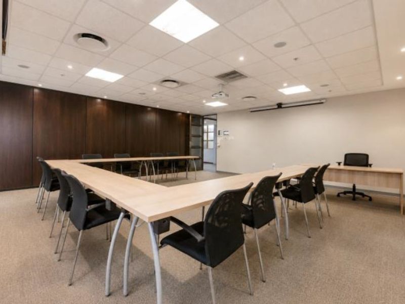 A professional commercial boardroom featuring wood paneling, carpeted flooring, a suspended ceiling with recessed lighting, and an integrated projector system.
