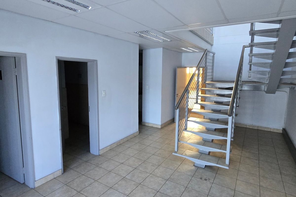 A spacious industrial hallway featuring tiled flooring, a modern metal staircase with stainless steel handrails, and a suspended ceiling with recessed lighting.