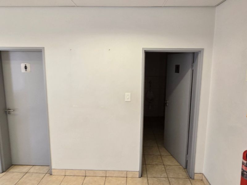 A functional industrial hallway featuring tiled flooring, drop ceiling, and wall-mounted fire safety equipment providing access to restroom facilities.