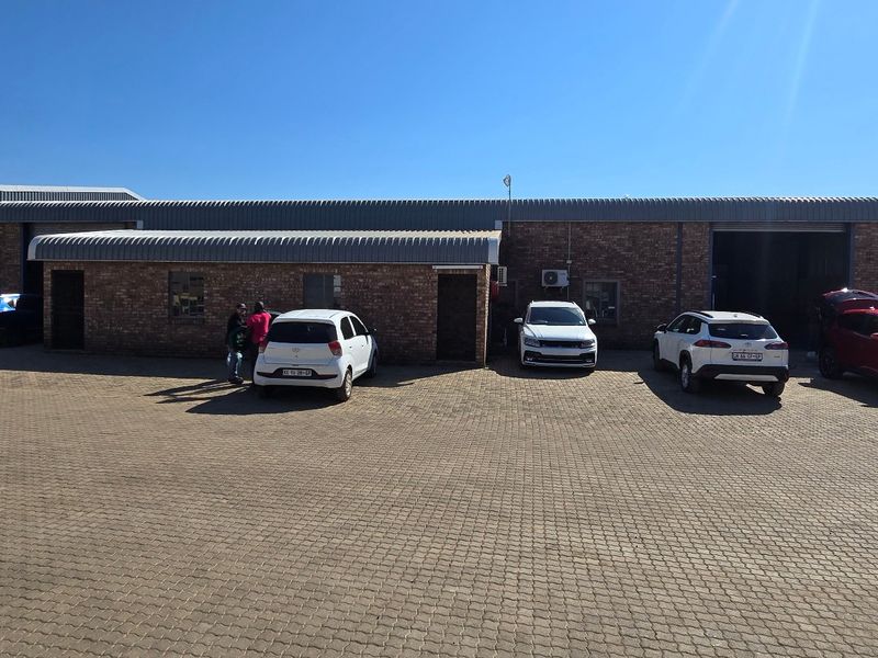 Industrial property exterior featuring a brick facade, corrugated metal roofing, and multiple roller shutter doors with a large paved parking area.
