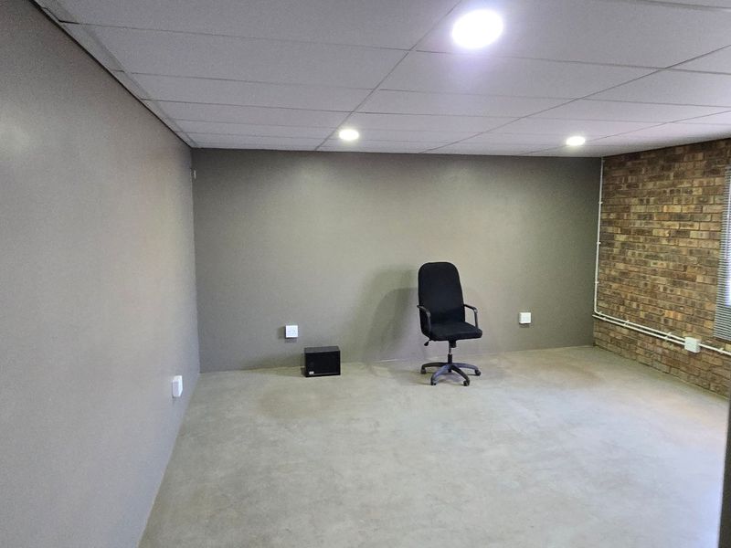 A clean, open-plan office space featuring a textured exposed brick wall, neutral painted walls, a suspended ceiling with recessed lighting, and durable concrete flooring.