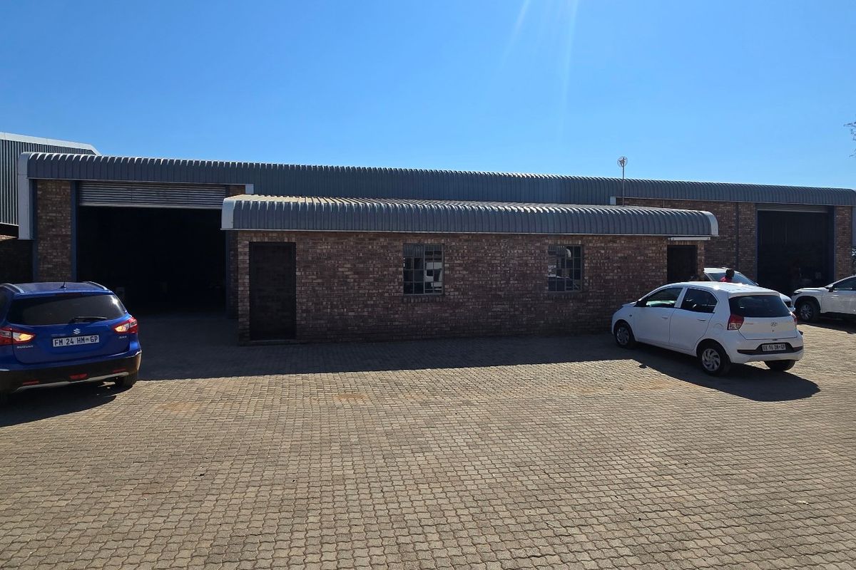 Front view of an industrial warehouse property featuring face brick construction, corrugated metal roofing, and multiple roller shutter doors with a paved yard area.